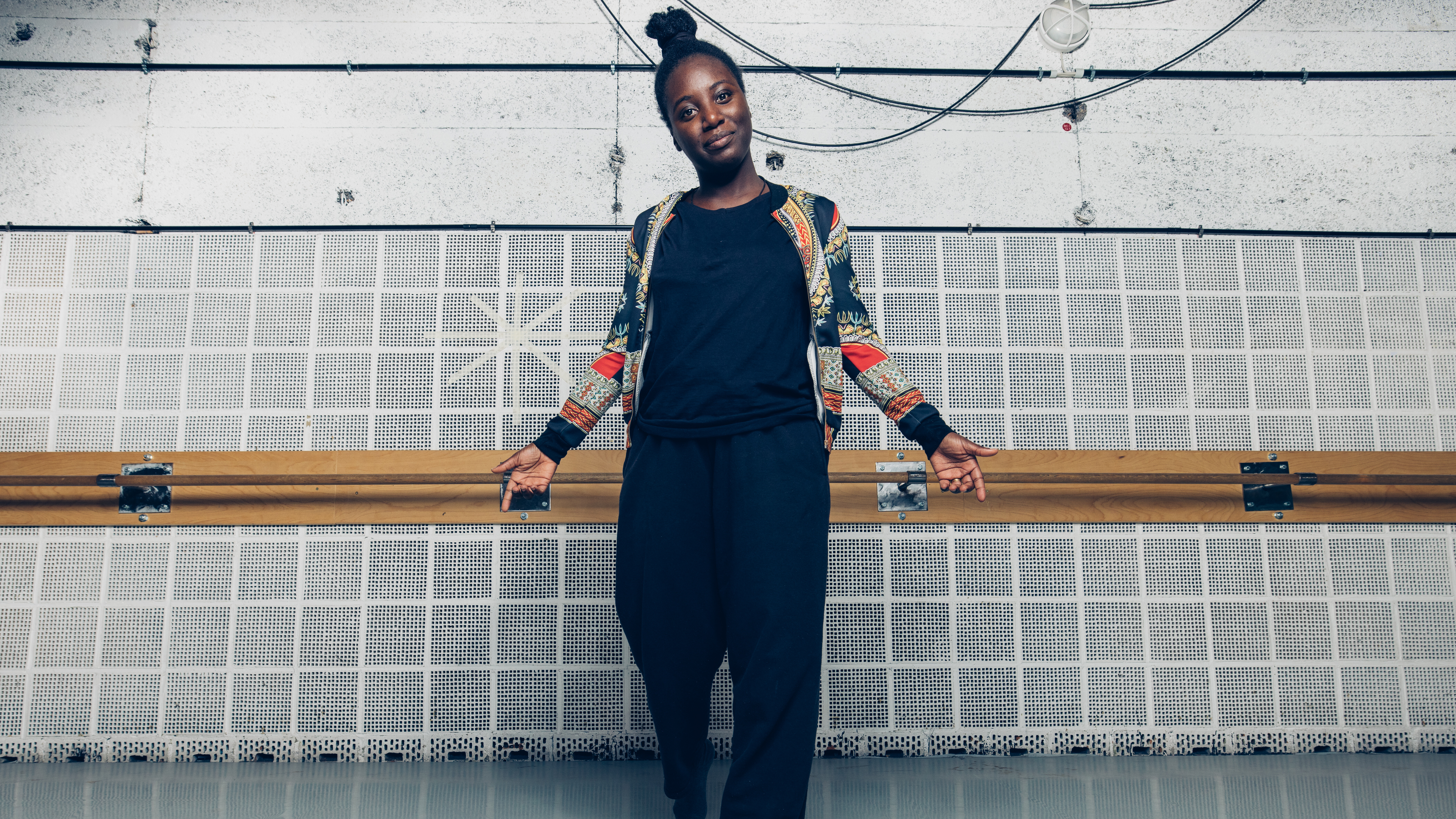 Ganzkörper-Bild von Jenny vor einer weiß-schwarz gemusterten Wand. Sie trägt einen schwarzen Einteiler und eine bunte Jacke und blickt in die Kamera.