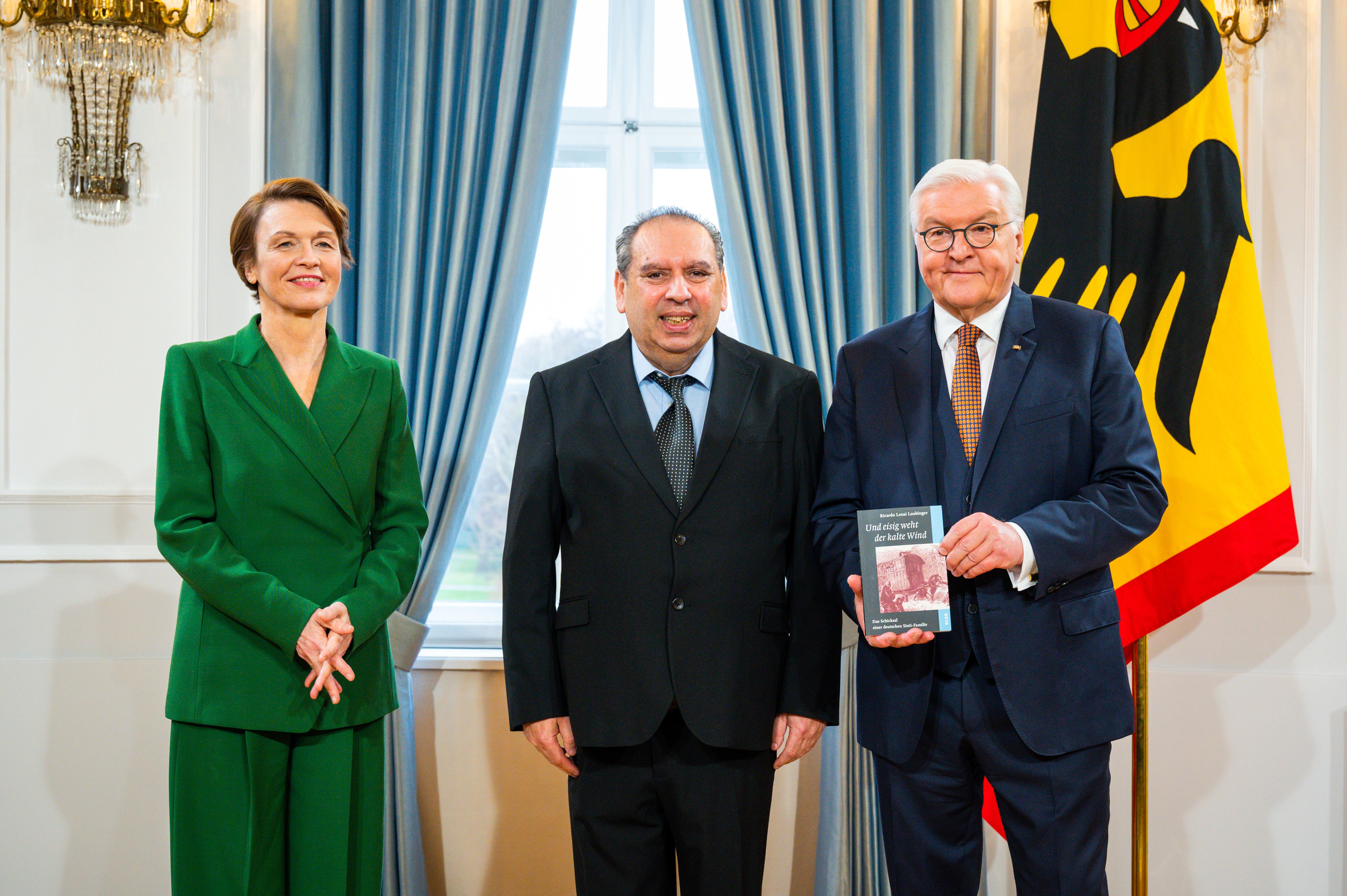 Ricardo Lenzi Laubinger steht im Anzug vor einer Fahne mit dem Bundesadler. Rechts von ihm steht Bundespräsident Frank-Walter Steinmeier. Er hält das Buch von Laubinger in der Hand. Links von Laubinger steht Elke Büdenbender, die Frau des Bundespräsidenten.