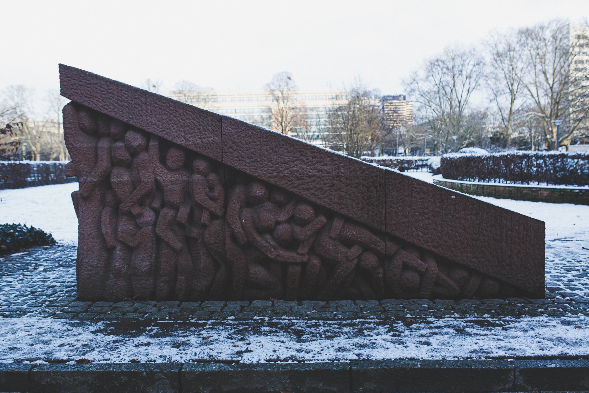 Auf einer schneebedeckten Wiese steht ein rot-brauner Sandsteinblock mit stilisierten Figuren im Hochrelief.