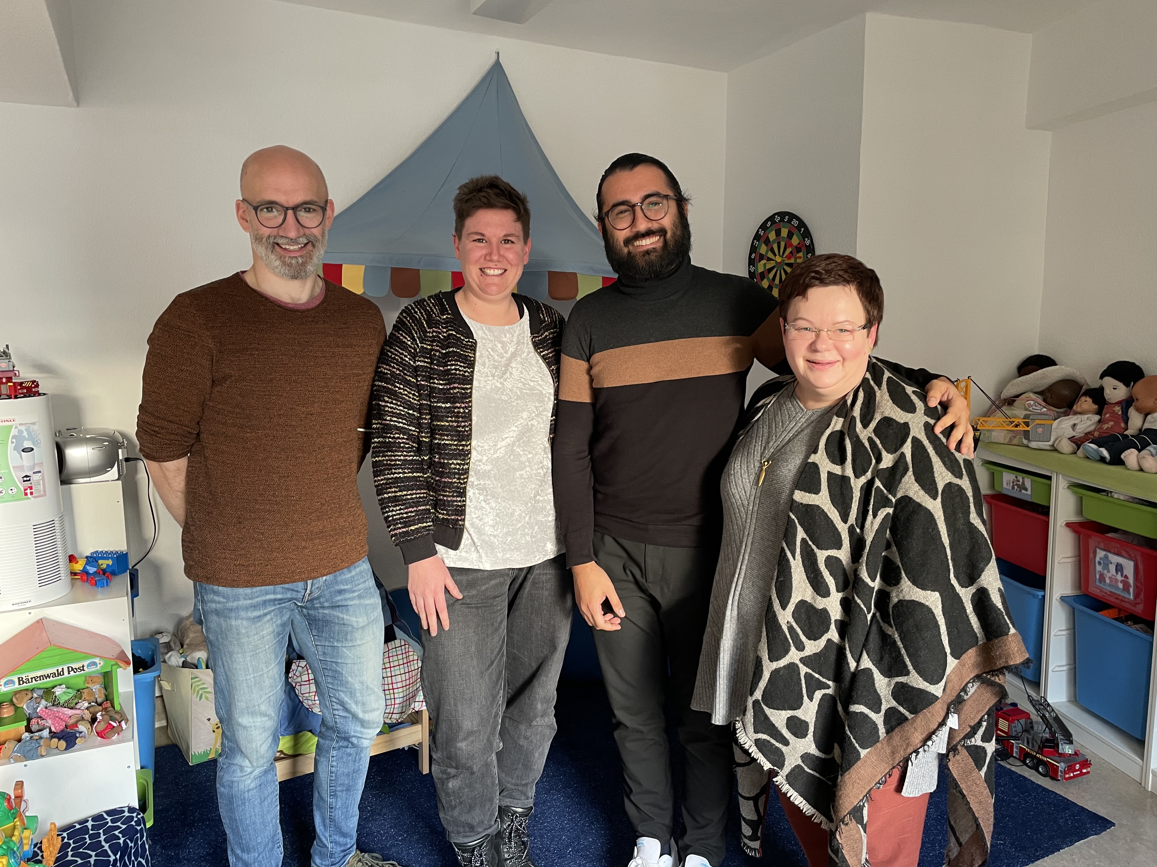 Vier Personen stehen eng nebeneinander in einem Raum und lächeln in die Kamera. Im Hintergrund stehen Regale mit Spielsachen.