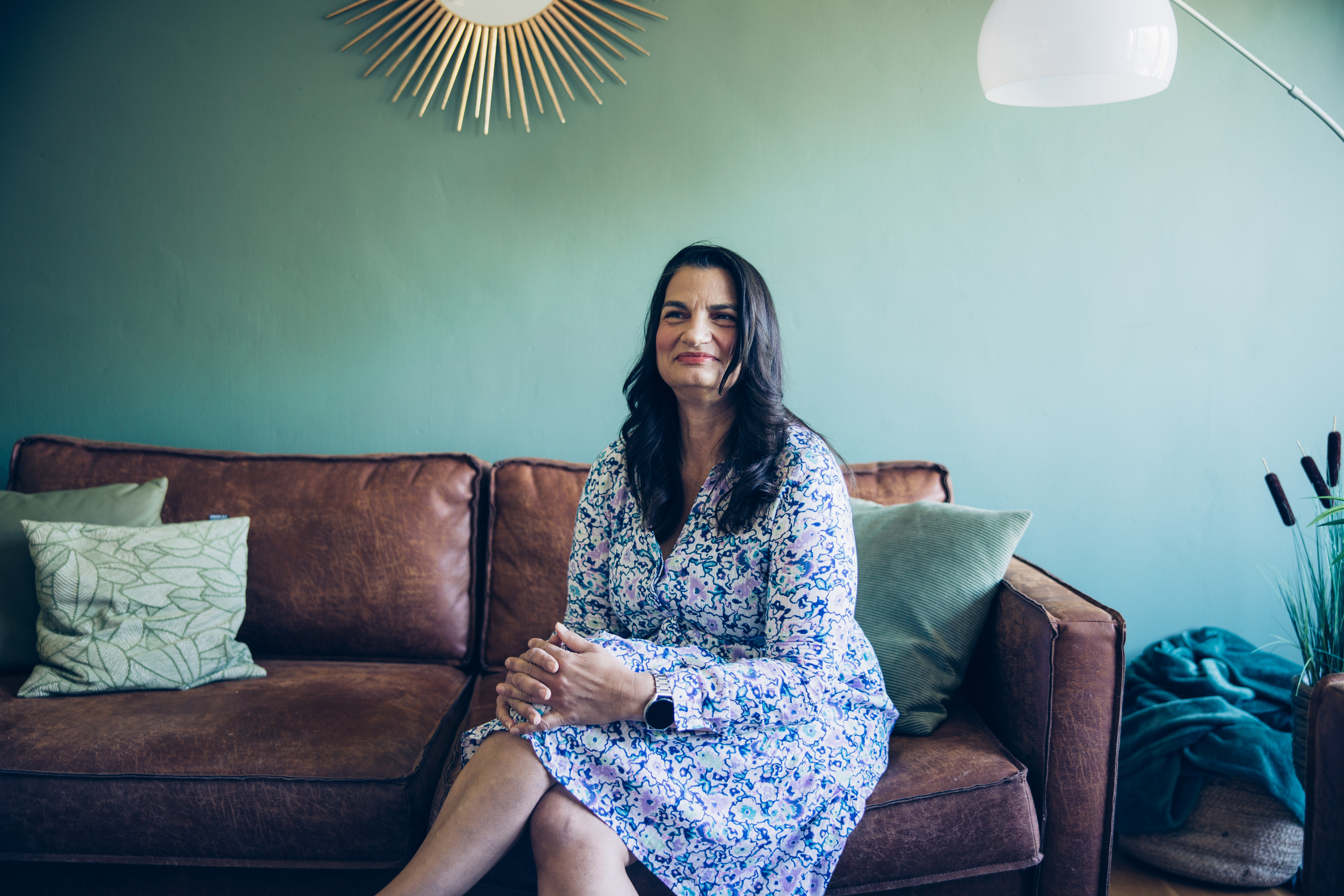 Eine Frau mit langen lockigen schwarzen Haaren. Sie sitzt in einem gemütlichen Wohnzimmer auf dem Sofa und lächelt selbstbewusst.