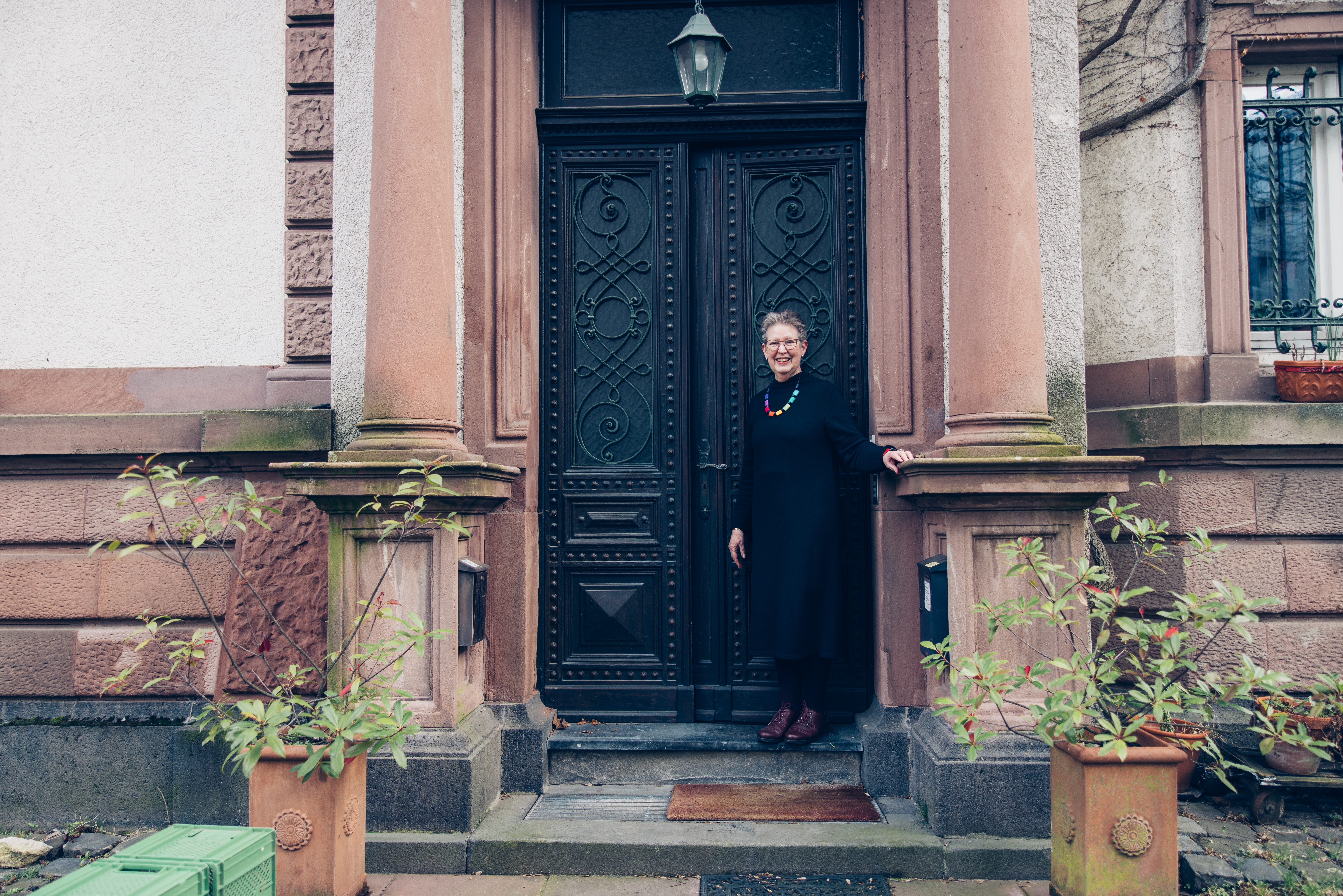 Eine ältere Frau steht vor dem Eingang einer denkmalgeschützten Villa und blickt Richtung Kamera. Sie lehnt ihre Hand an die Eingangssäulen.